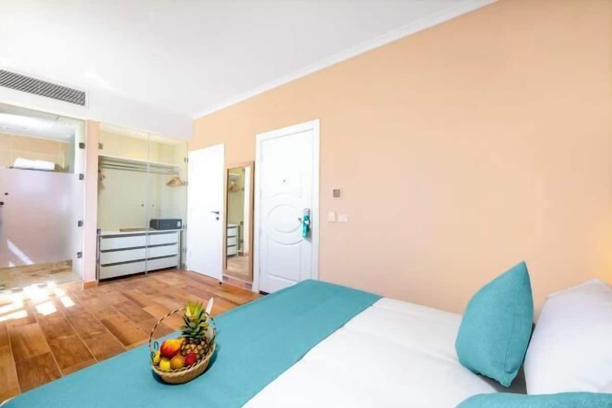 Une chambre d’hôtel avec un lit, un panier de fruits, un miroir et une porte donnant sur une salle de bain.