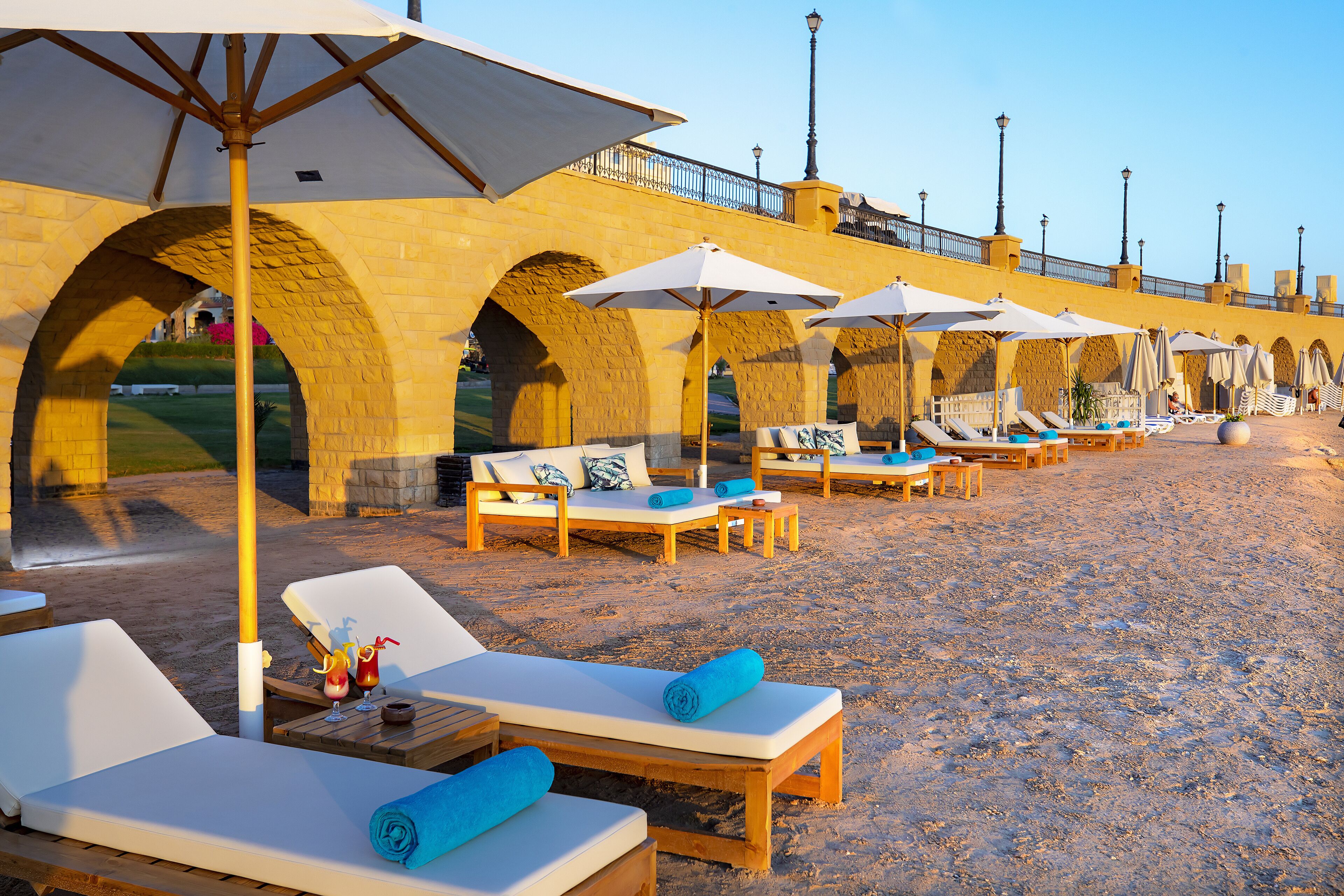 Een loungeruimte aan het strand met ligbedden, parasols en een stenen brug op de achtergrond.