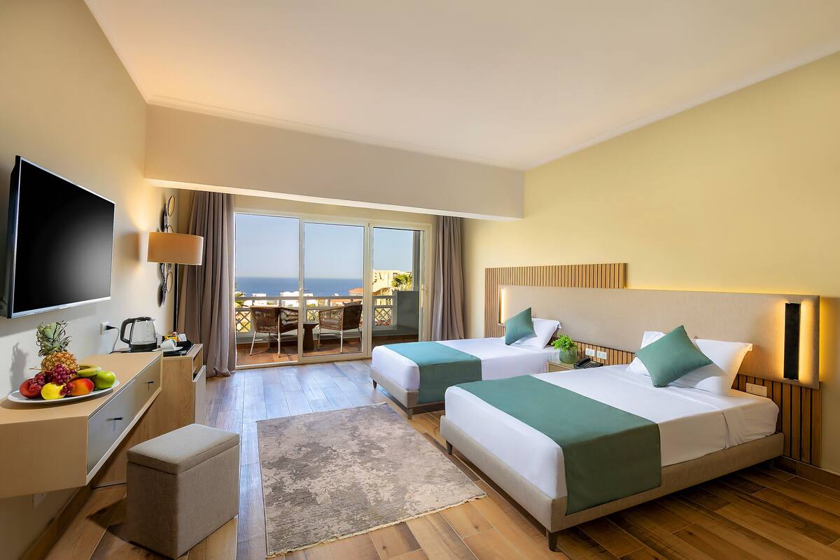 Une chambre d’hôtel avec deux lits, une télévision, un bureau avec un panier de fruits et un balcon avec vue.