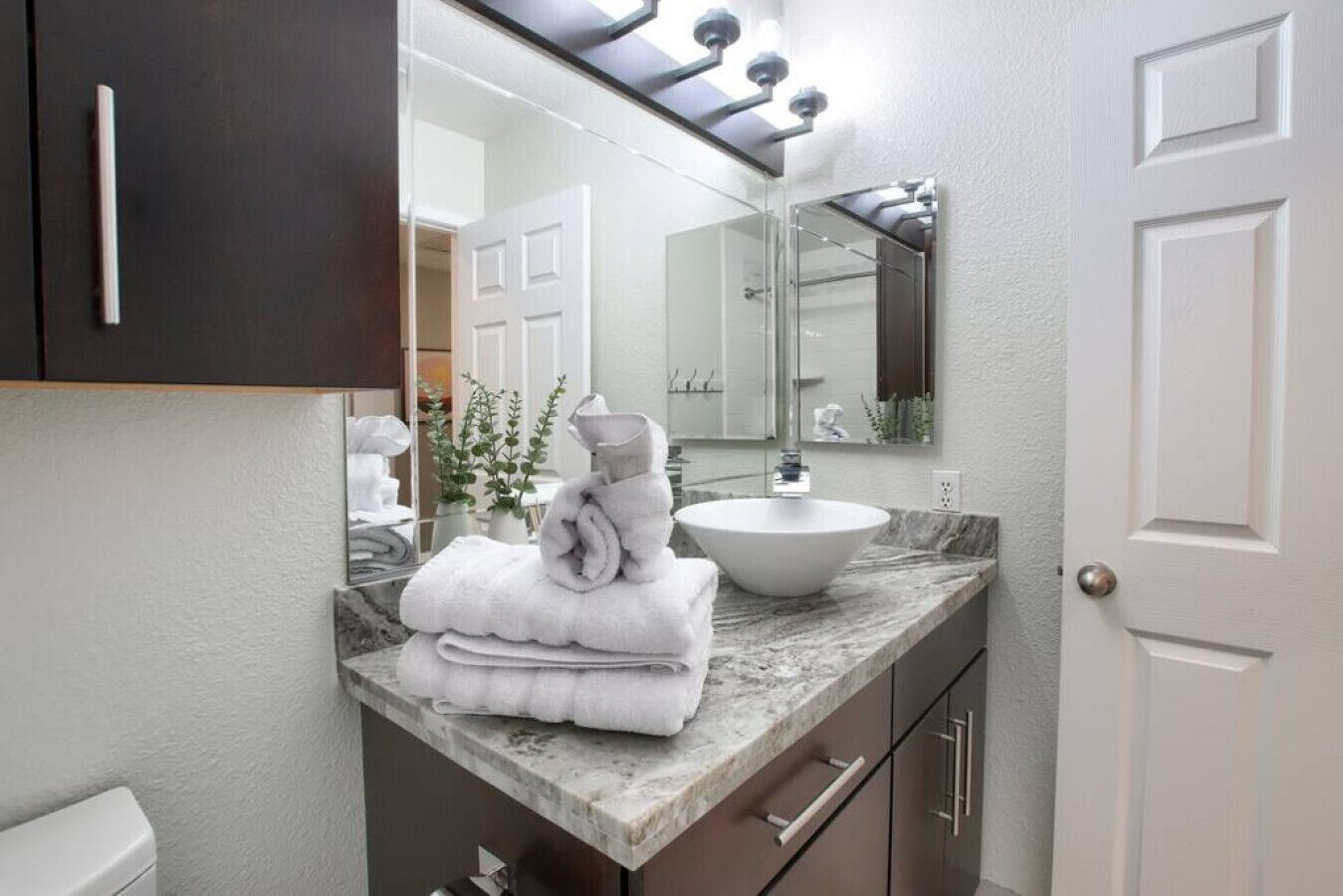 A bathroom with a marble countertop, a white sink, and a mirror above it. There are towels stacked on the countertop and a potted plant beside the mirror.