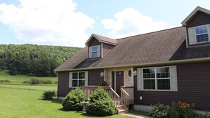 A two-story house with a front porch, a small garden, and a view of a forested hill. - Drive It Home Recent New Construction, 5 minutes to Dreams Park or Cooperstown (Cooperstown)