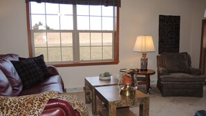 A living room with a leather sofa, a patterned armchair, a small table with a lamp, and a window with a view of a grassy area. - Drive It Home Recent New Construction, 5 minutes to Dreams Park or Cooperstown (Cooperstown)