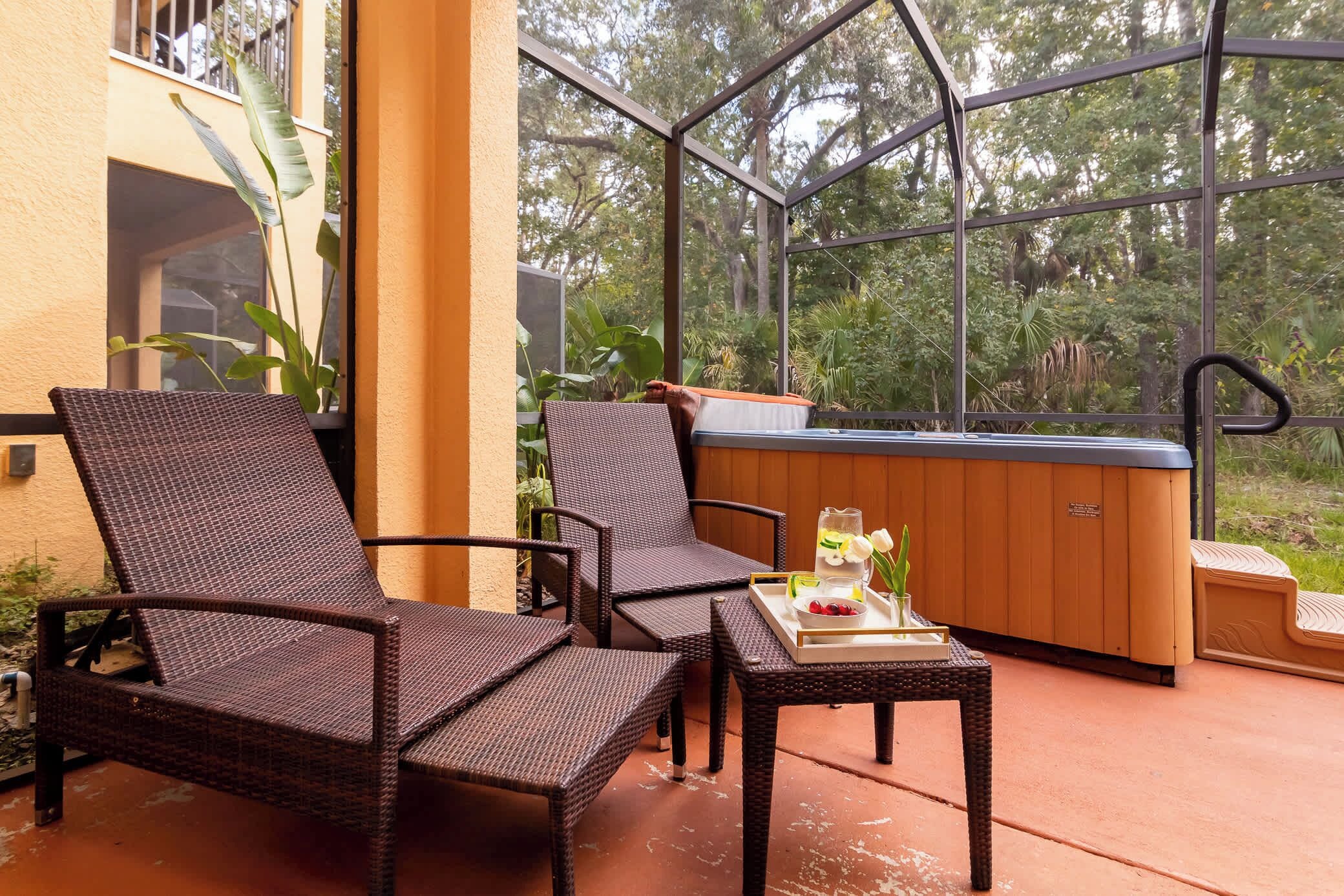 A glass-enclosed patio with wicker chairs, a wooden table with a tray of fruit and a drink, and a hot tub.