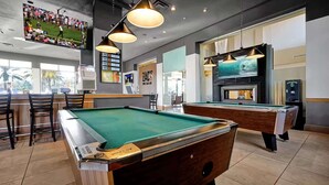 A pool table in a room with a bar area, a television displaying a sports event, and a fireplace.