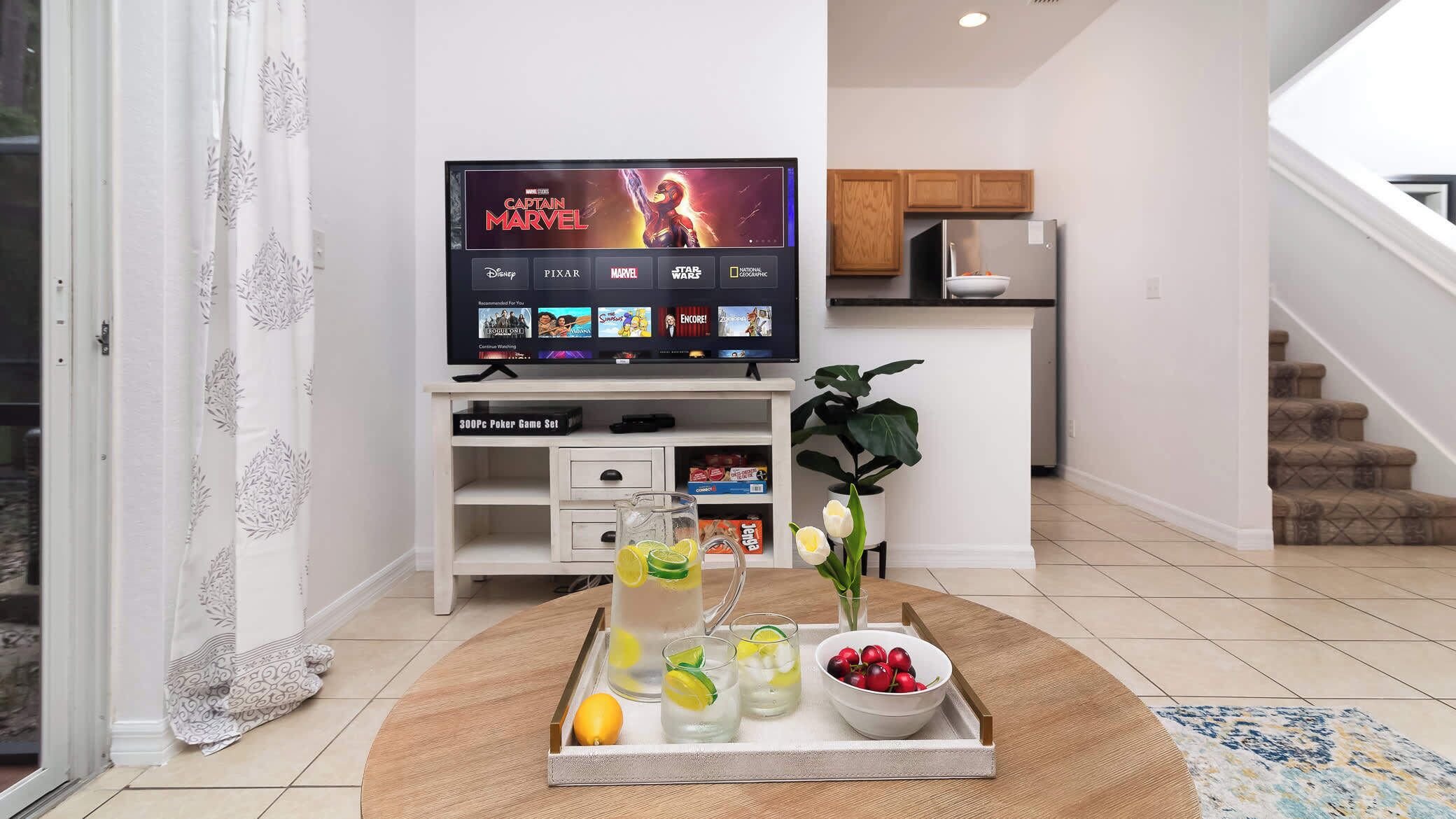 A living room with a flat-screen TV displaying a movie, a coffee table with drinks and fruit, and a staircase in the background.