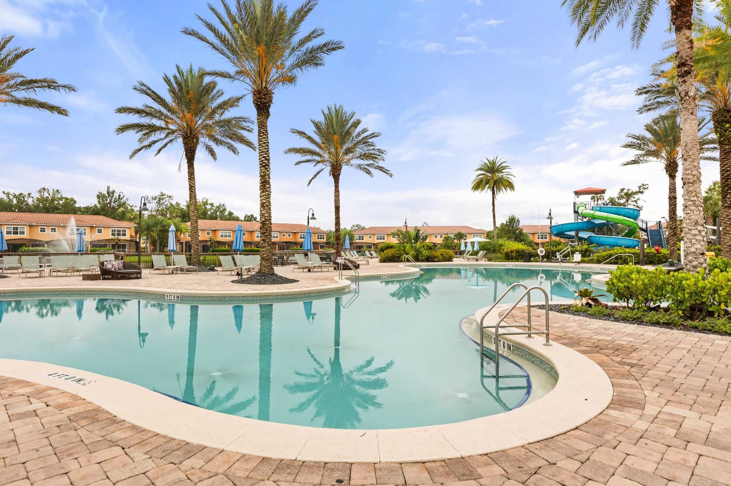 A swimming pool with a water slide, palm trees, lounge chairs, and a brick patio.