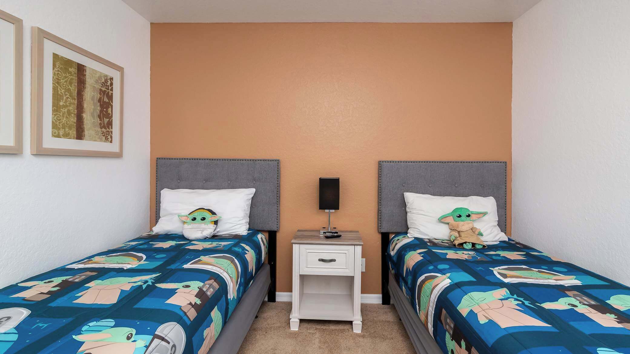 A hotel room with two beds, each with a headboard and a pillow featuring a character. There is a bedside table with a lamp and a drawer.