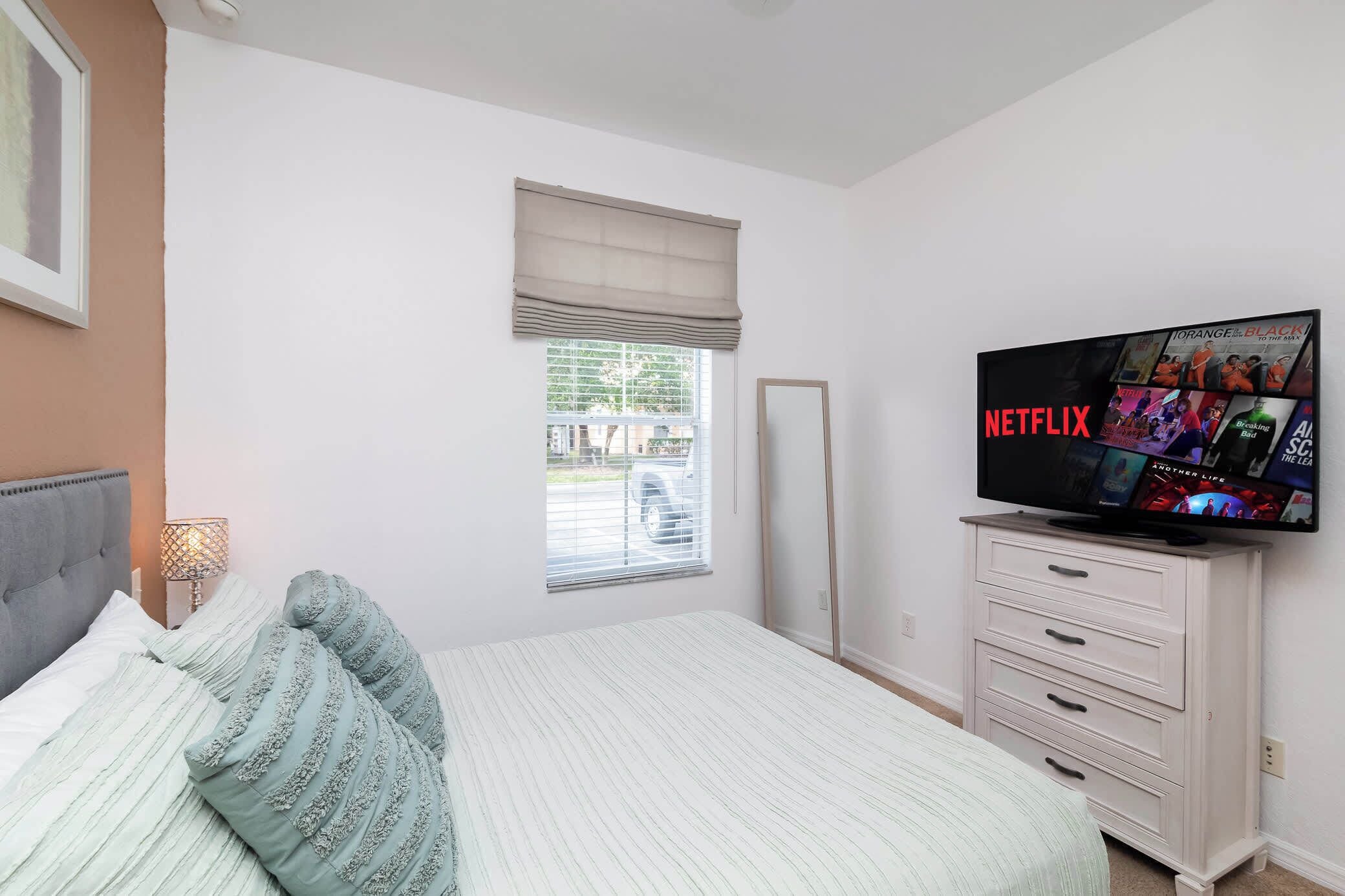 Een slaapkamer met een bed, een televisie op een dressoir, een raam met jaloezieën en een lijst met een afbeelding aan de muur.