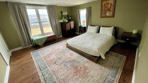 A bedroom with a large bed, a wooden floor, a patterned rug, a window with curtains, a nightstand, and a painting on the wall.