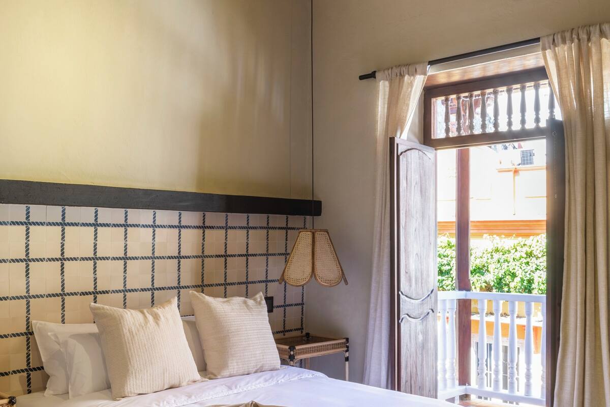 A bedroom with a bed, a nightstand, a lamp, and a view of the outside.