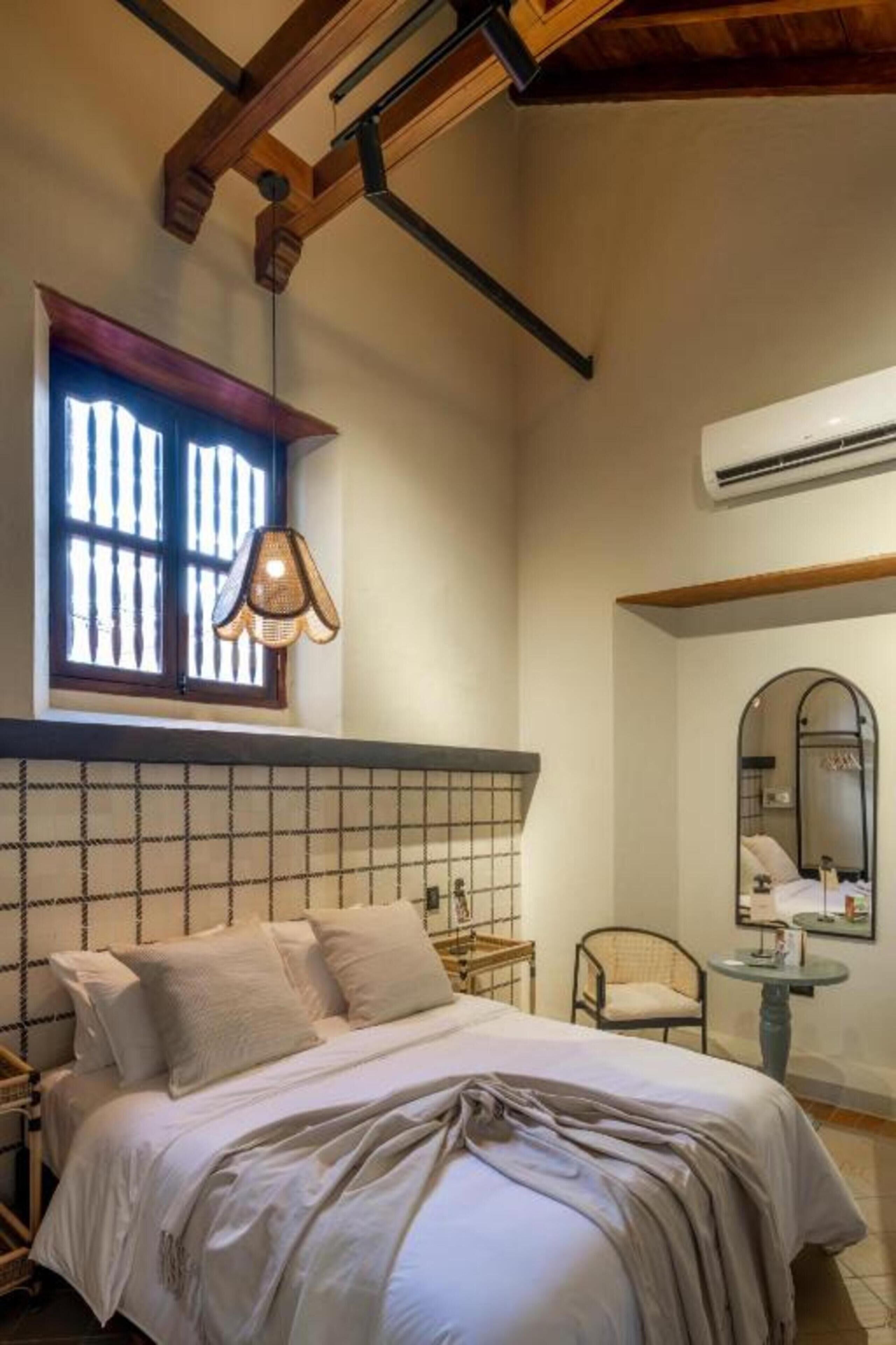 A bedroom with a bed, a window with a hanging light, a mirror, and a small table.
