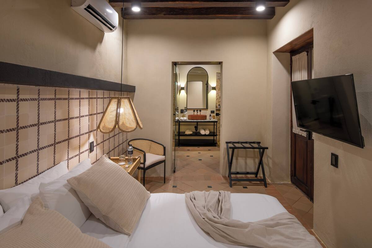 A hotel room with a bed, a chair, a small table, a TV, and a bathroom visible through an arched doorway.