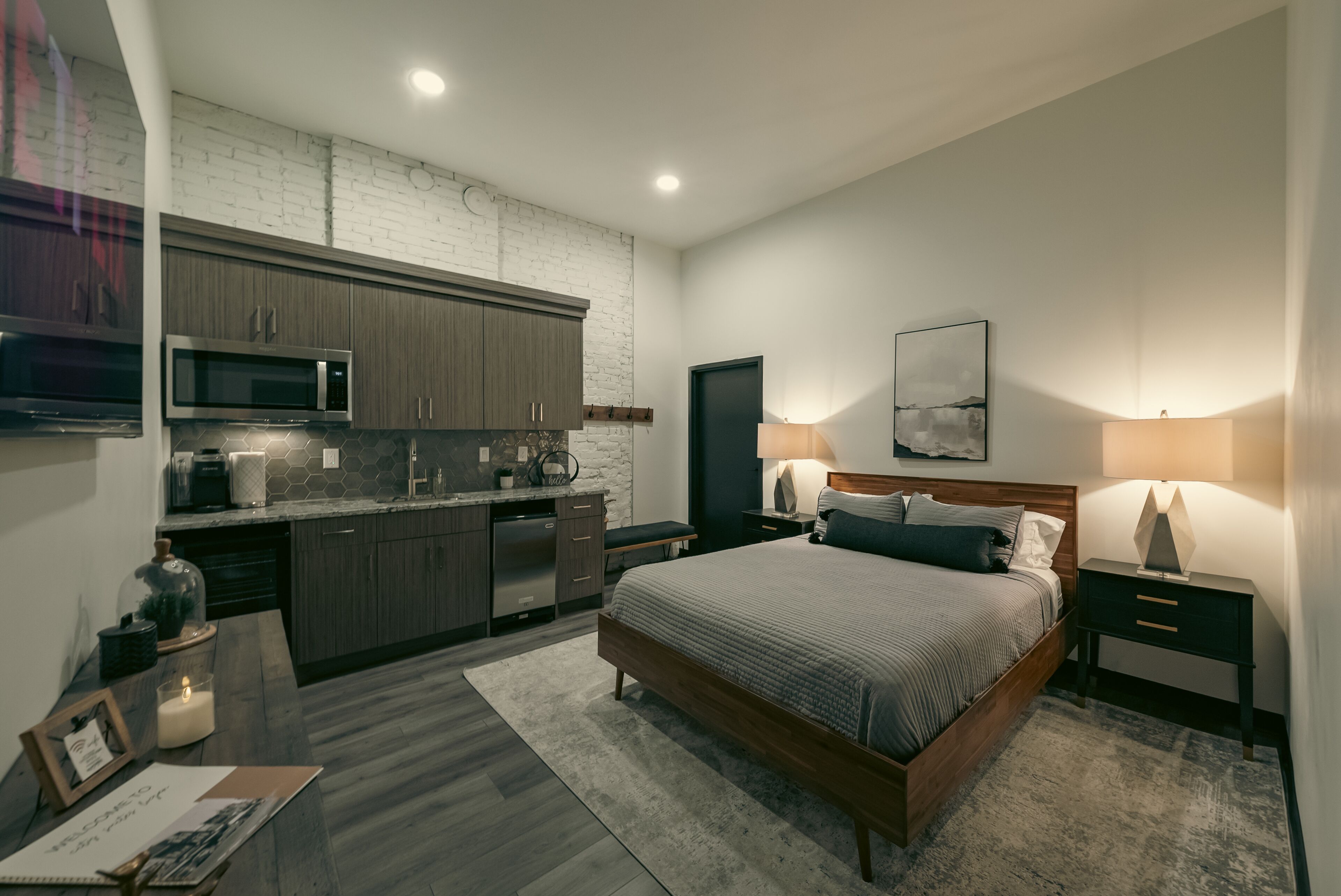 Een moderne slaapkamer met een groot bed, nachtkastjes, een bureau en een kitchenette met een magnetron en kasten.