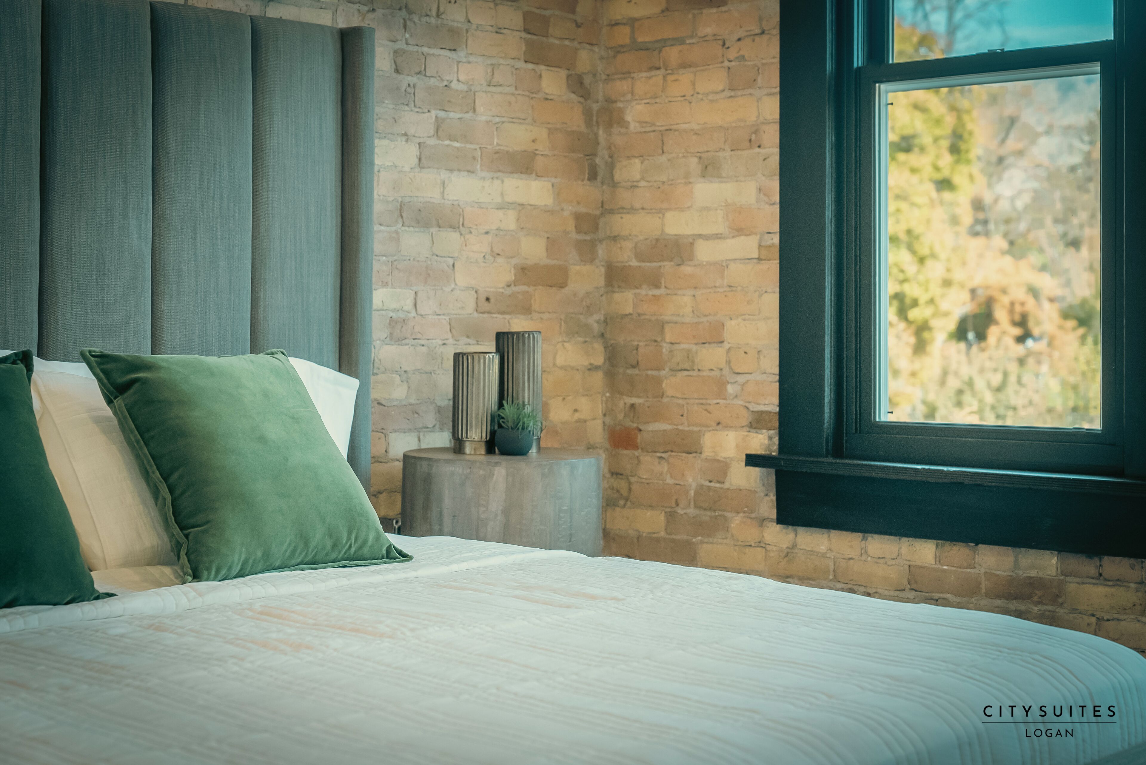 Une chambre avec un mur en briques, une grande fenêtre et un lit avec une literie verte et blanche.