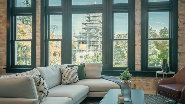 A modern living room with a beige sectional sofa, a coffee table, and a red armchair. Large windows offer a view of a construction site outside. - Luxury Suite ~ Above Starbucks ~ Main St View (Logan)