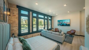 A modern bedroom with a large bed, a sofa, and a chair, large windows with a view, and a painting on the wall. - Luxury Suite ~ Above Starbucks ~ Main St View (Logan)