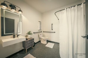 A modern bathroom with a white sink, mirror, and toilet. There are black floor tiles, a white shower curtain, and a black floor lamp. - Luxury Suite ~ Above Starbucks ~ Main St View (Logan)