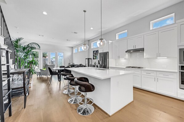 A modern kitchen with white cabinetry, a central island with bar stools, stainless steel appliances, and pendant lighting. - Modern Home near Medical Center (Houston)