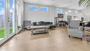 Un salon spacieux avec un canapé gris, une table basse et un fauteuil à motifs. De grandes fenêtres offrent une vue sur la verdure environnante.