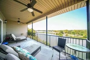 Een overdekt balkon met een plafondventilator, een bank, een tafel en uitzicht op een rivier en bomen.