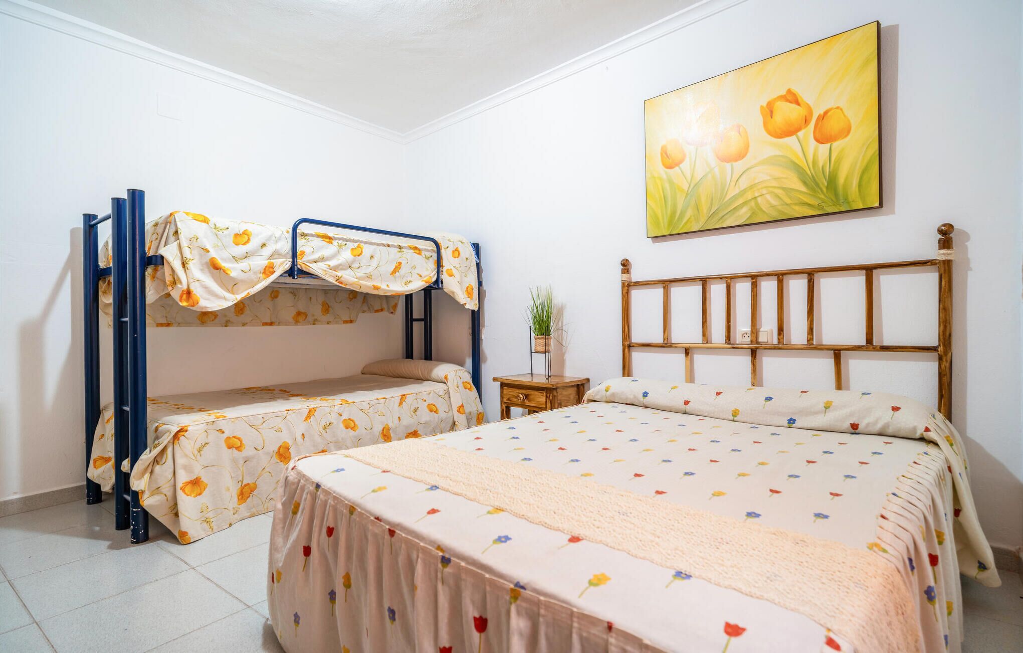 A bedroom with bunk beds, a single bed, a wooden headboard, a small bedside table, and a wall-mounted painting of flowers.