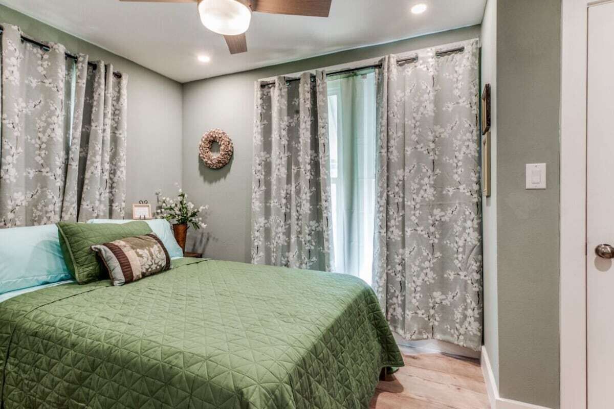 Un dormitorio con colcha verde acolchada, cortinas florales, ventilador de techo de madera y una pequeña planta en maceta.