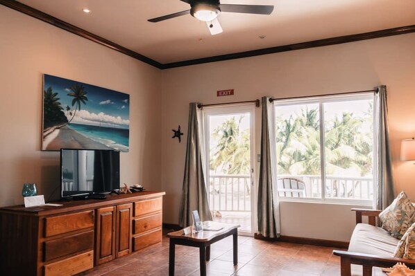 Una stanza con una TV su un mobile in legno, un quadro raffigurante una spiaggia, un segnale di uscita, un balcone con tende e un divano.