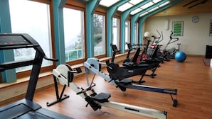 A gym with multiple cardio machines, large windows, and a wooden floor.