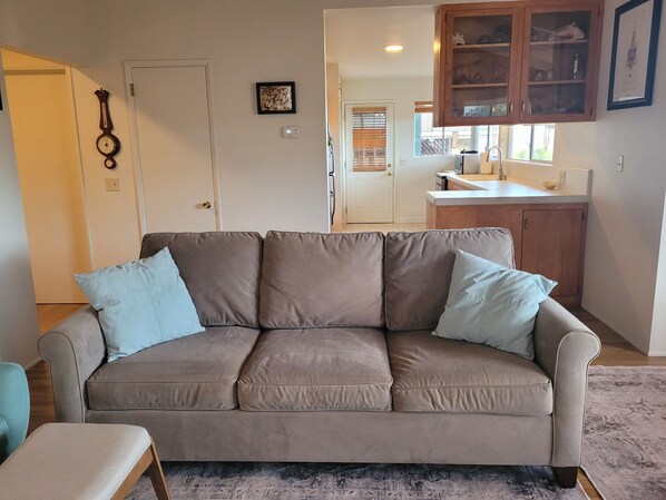 Una sala de estar con un sofá color beige, una mesita y una cocina visible a través de una puerta abierta.