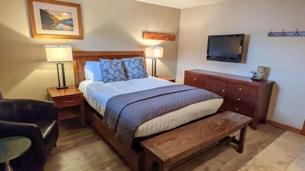 Un dormitorio ordenado con una cama, lámparas de noche, un banco de madera, un televisor montado en la pared y un perchero.
