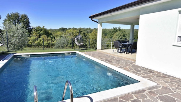 Une piscine avec une terrasse en pierre, un patio couvert agrémenté de mobilier d’extérieur, et une vue sur les arbres et les collines en arrière-plan.