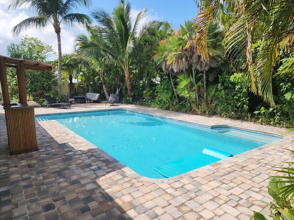A rectangular swimming pool with a tiled edge, surrounded by lush greenery and palm trees. - Thumb Point Oasis w/Heated Pool- walk to beach! (Fort Pierce)