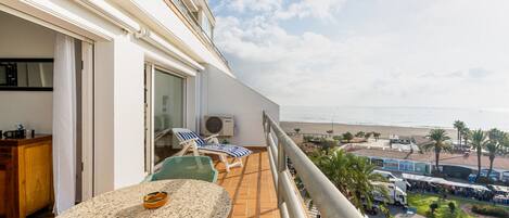 Un balcón con mesa y sillas, con vista a una playa y un pueblo con tiendas y palmeras.