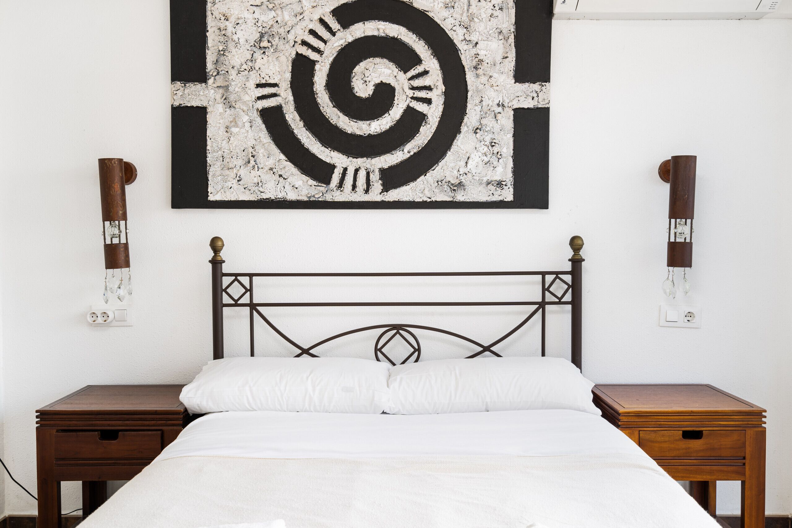 A bedroom with a bed, two bedside tables, a wall-mounted artwork, and a wall-mounted light fixture.