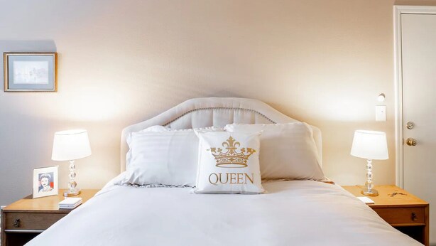 Een net opgemaakt bed met witte beddengoed en een kussen met een kroon en de tekst “QUEEN”. Twee nachtkastjes met lampen en een ingelijst foto op een ervan.