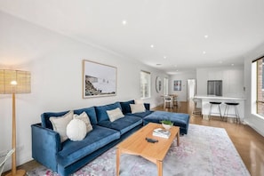 Ein modernes Wohnzimmer mit einem blauen Sofa, einem Holztisch und einer Küchenzeile im Hintergrund.