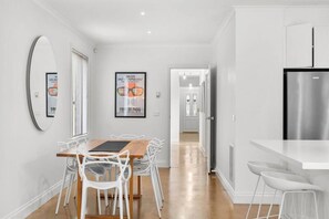 Une cuisine moderne avec un réfrigérateur en acier inoxydable, un coin repas avec des chaises blanches et une table en bois, et un couloir menant à une porte.