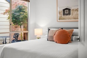 Ein Hotelzimmer mit einem Bett, einer Lampe, einem kleinen Tisch und Blick auf einen Baum und ein Gebäude durch das Fenster.
