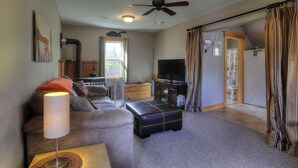 Ein Wohnzimmer mit Sofa, Hocker, Stehlampe, Deckenventilator, Fernseher und Blick in einen anderen Raum durch eine Tür.