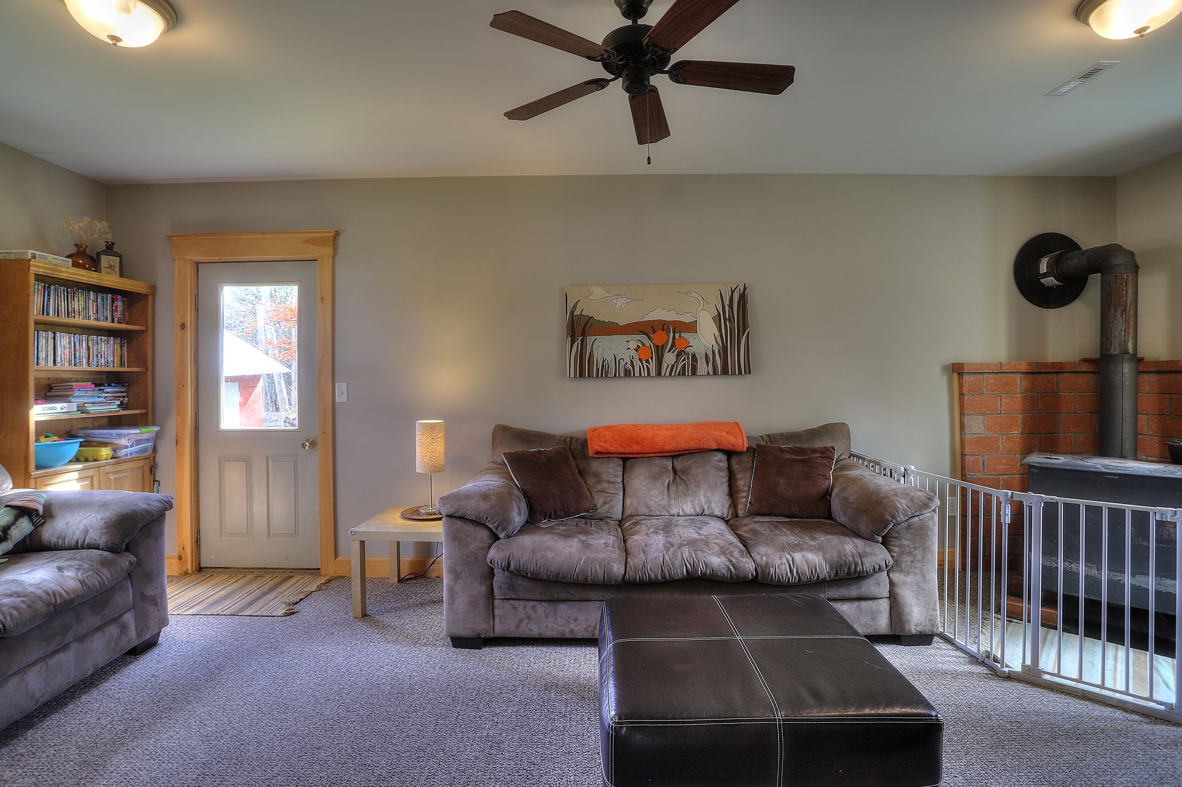 A cozy living room with a fireplace, a ceiling fan, a bookshelf, and a door leading outside.