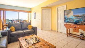 A living room with a sofa, a coffee table with a chessboard, a television, and a view of a cityscape through large windows. - Amazing 3BR 2Bath at the Princesa Resort A-505 Ocean View Condo (Puerto Peñasco)