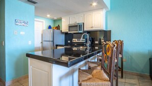 A kitchen with white cabinets, a black countertop, a microwave, a refrigerator, and a sink. There is a wooden chair with a sunflower design. - Amazing 3BR 2Bath at the Princesa Resort A-505 Ocean View Condo (Puerto Peñasco)