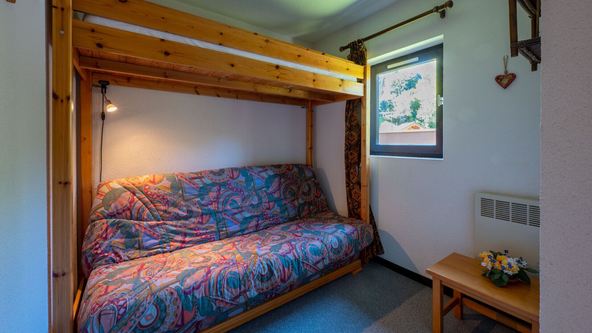 Ein gemütliches Zimmer mit einem farbenfrohen gemusterten Sofa, einer Holzbalkendecke, einem kleinen Fenster mit Aussicht und einer Holzbank mit Blumen.