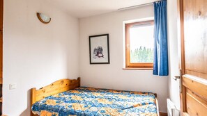 Une petite pièce avec un lit, une fenêtre donnant sur des arbres, un tableau encadré au mur et une porte en bois.