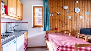 Une petite cuisine équipée d’une machine à café, d’un évier et d’une cuisinière. On y trouve un lit recouvert d’une nappe à carreaux et une fenêtre avec des rideaux.
