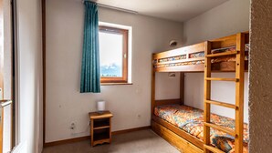 A small, cozy room with a bunk bed, a window with curtains, a bedside table, and a view of mountains.