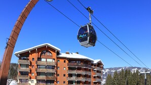A cable car system with a wooden structure and a building with multiple balconies and a ski slope in the background.