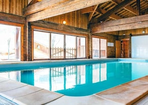 An indoor swimming pool with wooden beams, large windows, and a tiled floor.