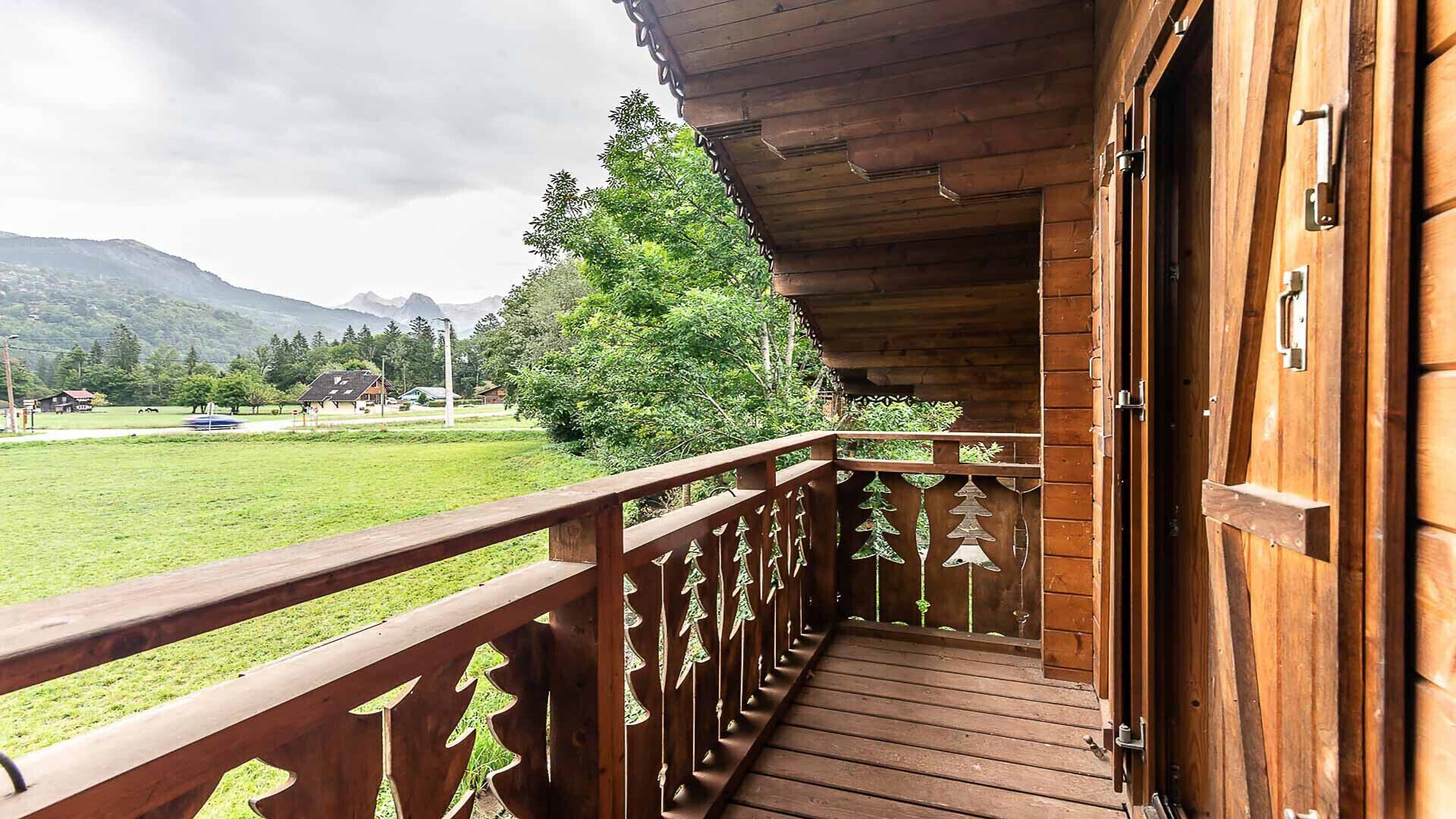 Een houten balkon met een leuning, een deur en uitzicht op een groen veld en bergen.