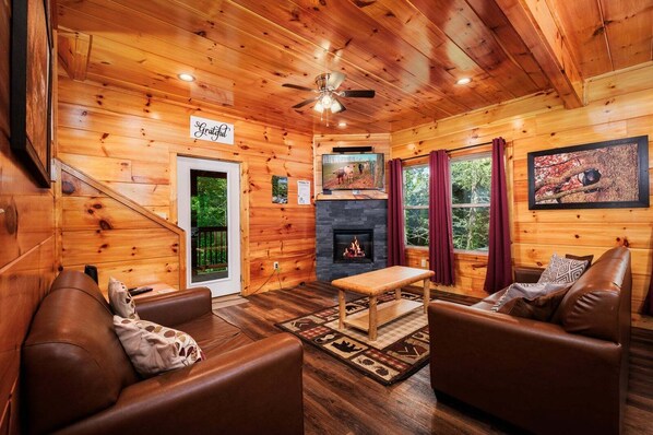 Interior acogedor de una cabaña con paredes de madera, chimenea, sofás de cuero y un ventilador de techo.